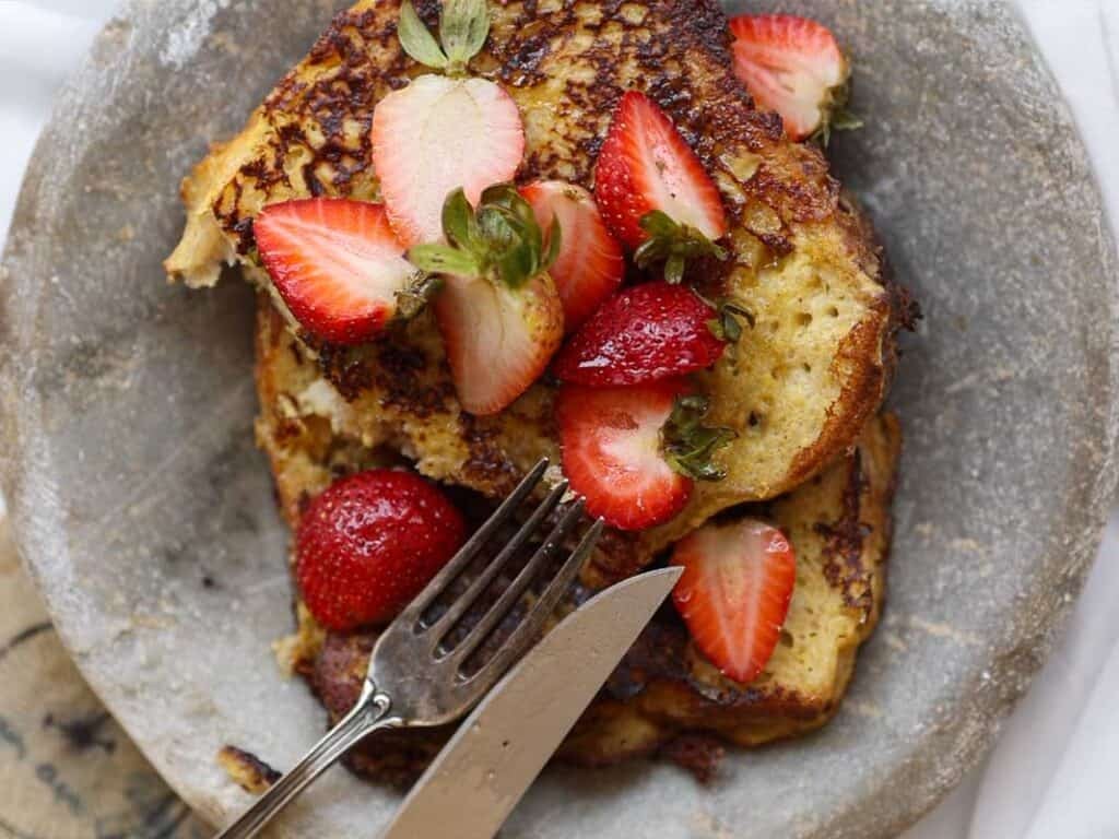 A plate of vegan French toast topped with sliced fresh strawberries. A fork and knife rest on the plate, ready to cut into the toast. The toast appears golden brown and crispy.